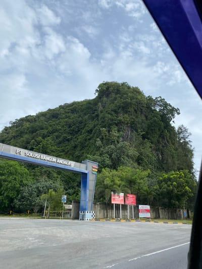 A landscape of Naga Umbang, Indonesia, features a verdant, densely wooded hillside alongside a gray asphalt road. A prominent blue and silver structure, likely a company entrance, stands near the road. The sky is overcast with scattered white clouds. The scene is partially viewed from within a vehicle, as indicated by the blue structure in the upper left corner. Several signs are posted along the base of the hillside, displaying red and white informational notices.
