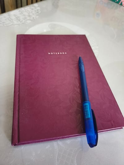 A close-up, high-angle shot shows a closed, dark pink notebook with a textured floral pattern on its cover. The word "NOTEBOOK" is embossed in silver lettering on the upper right side of the cover. A bright blue ballpoint pen with a silver clip rests diagonally across the notebook, with its tip pointing towards the upper left. The pen has a ribbed grip section and a barcode sticker on its barrel. The notebook and pen are placed on a white tablecloth with a subtle, embossed pattern. The background is blurred, but suggests a domestic setting. There is no indication of location within Podkletnoe, Russia.