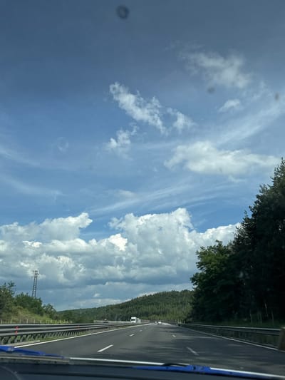 The image is a view from a car driving on a highway. The sky is blue with scattered white clouds, some of which are large and puffy, while others are thin and wispy. There are some dark circular spots on the sky, likely due to a dirty lens. On either side of the highway, there are green trees and hills. A power line pylon is visible on the left side of the road. There are other vehicles on the highway in the distance, including a white truck. The road is asphalt and has lane markings. The photo appears to have been taken in Krushovitsa, Bulgaria, a rural area with lush greenery and rolling hills.