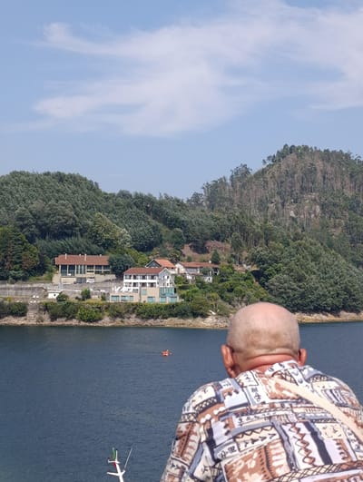 The image depicts a scenic view of a body of water in Terras de Bouro, Portugal, with a verdant hillside dotted with trees and buildings in the background. In the foreground, from a rear view, a bald man wearing a patterned shirt looks out at the landscape. The water appears calm, with a small orange raft carrying two individuals visible in the distance. The hillside features a complex of modern buildings with terracotta roofs and large windows, suggesting a residential or commercial development. The overall impression is one of tranquility and natural beauty, enhanced by the presence of human activity in the form of the distant raft and the buildings.