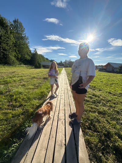 In the city of Zamosh'e (Zaklinskoe S/P), Russia, on a sunny day, two young girls and a dog are walking on a wooden boardwalk. The boardwalk is surrounded by lush green grass and trees. In the background, there are several glamping tents. The sun is shining brightly, casting long shadows. The girl in the foreground is wearing black shorts and a white t-shirt, with a walkie-talkie tucked into her shorts. The other girl is wearing a tie-dye t-shirt and denim shorts, holding a walkie-talkie in her hand. The dog, a Cavalier King Charles Spaniel, is walking ahead of them.