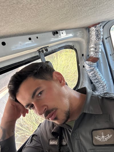 A male individual is depicted in a portrait-oriented image taken inside a vehicle, located in Beit Horon, Palestinian Territory. The subject, positioned in the foreground and viewed from the chest up, has dark hair and a trimmed beard, looking directly at the camera with a neutral expression. His left hand is raised, supporting his head. He wears a grey collared uniform shirt, which features an emblem of the Israel Prison Service (IPS) on the right sleeve and a name tag in Hebrew, "מאתם" (Matam), on the left chest area. A tattoo is visible on his left forearm. The vehicle's interior consists of a light-colored headliner and exposed light grey painted metal framing around a window. Along the right-side pillar of the vehicle, a corrugated metallic foil duct, possibly an improvised air vent, runs vertically, secured with wire. Through the vehicle window, a dense outdoor environment with tall, green vegetation is visible under bright lighting conditions.
