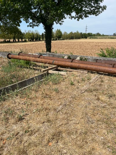The image depicts a rural scene in Castiglione delle Stiviere, Italy, under a clear blue sky. In the foreground, dry, golden grass covers the ground. Several large, rusty metal pipes are laid out horizontally. These pipes rest on a wooden support structure. Behind the pipes, a low concrete structure runs horizontally, partially obscured by vegetation. Beyond this, a large field of tilled, brown earth stretches into the distance. A line of trees borders the field, with more distant trees and vegetation visible further back. A power pylon and electrical lines are visible on the horizon. The scene is one of an outdoor, agricultural or industrial landscape. There are no people or animals visible in the image.