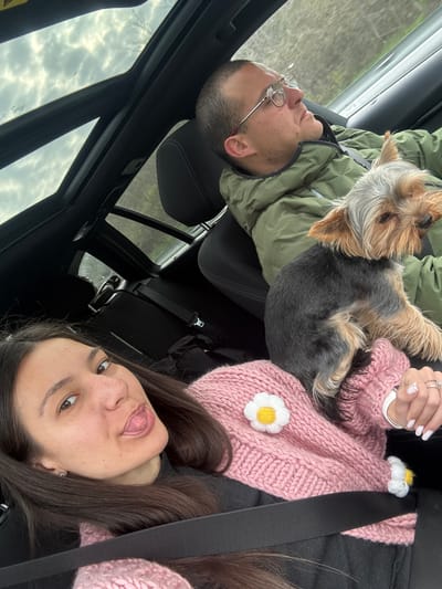 A selfie was taken inside a car showing two people and a dog. The man is on the left wearing glasses and a green jacket. A Yorkshire Terrier dog is on top of him. The woman is on the right, wearing a pink sweater with flower appliques and a seatbelt. The car has a sunroof with a view of cloudy sky. There is no evidence to suggest the location is in Kipra, Bulgaria.