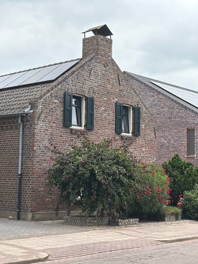 The image shows a brick house in Melick, Netherlands, with a roof covered in solar panels. The house has two windows with dark green shutters on the front facade. A chimney with a metal cap is visible at the top of the house. In front of the house, there is a large green bush with red flowers and two smaller evergreen bushes. The foreground consists of a paved sidewalk and road. The sky is overcast.