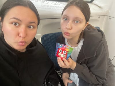 Two young women are seated on a train. The woman on the left is facing forward, looking at the camera with her lips pursed. The woman on the right is turned towards the left, holding a colorful candy packet with a cartoon monster on it. Both women are wearing casual clothing, and appear to be in a train car. The background shows seats and the interior of the train car. There is text visible on the wall behind them, which appears to be a train schedule or advertisement, possibly indicating they are in or near Рассудово, Russia.