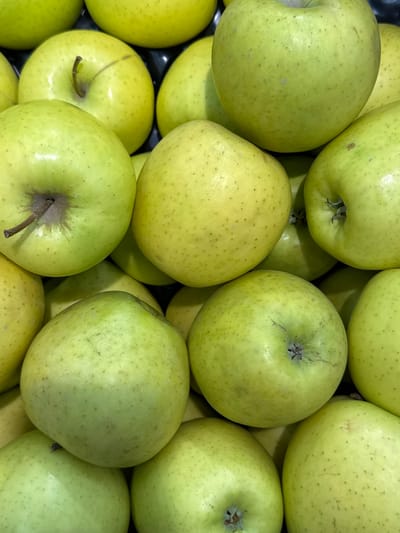The image displays a close-up, overhead view of a large pile of green apples. The apples are predominantly a light green color with subtle variations in shade and some appear to have small, dark specks on their skin. They are tightly packed together, obscuring the background which seems to be a dark surface, possibly a bin or crate. The lighting is bright, highlighting the smooth, rounded surfaces of the fruit. There are no people or any other objects visible in the frame. The scene appears to be a retail display of produce, likely in a market or grocery store. The location of Rozhdestvenskaya, Russia is not visually discernible from the image itself.