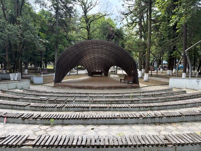 The image depicts an outdoor amphitheater in the city of Khoni, Georgia. In the foreground, curved rows of concrete steps are visible, some of which are partially covered with weathered wooden planks, suggesting a state of disrepair. A single red plastic bottle rests on the lower steps.

Dominating the mid-ground is a large, arched, open-air stage structure with a metal frame and a translucent covering. Benches are placed on the stage, and a single wooden bench with a metal frame is situated to the right of the stage.

The background is filled with lush greenery from a dense forest of tall trees, characteristic of Khoni's natural environment. The sky is overcast. No people are present in the scene. The overall impression is that of a public space in a park or garden setting.