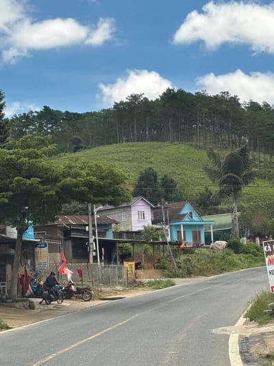 The image depicts a rural scene in Đưng K'Si, Vietnam, with a sloping, tree-covered hillside dominating the background under a blue sky with scattered white clouds. In the mid-ground, a paved road curves to the right, with a narrow sidewalk bordering it. Along the sidewalk, a series of buildings are visible.

On the left, a person in a dark hooded jacket is seated on a black motorcycle. To the right of the motorcycle, a red scooter is parked. A weathered building with a corrugated metal roof is situated behind the motorcycles, with various items displayed outside, including what appears to be clothing and bags. A sign with Vietnamese text is also visible on this building.

Further to the right, two distinct houses stand out: one is painted lavender with white trim and windows, and the other is a bright turquoise with white trim and pillars. Both houses have pitched roofs. Behind these houses, more buildings with green roofs and various trees and vegetation are present. A tall palm tree stands to the right of the turquoise house.

In the far right of the image, a portion of a large white sign with red text is visible, advertising "CA NHUNG" and "NU". The overall impression is one of a quiet, somewhat rustic settlement nestled at the base of a forested hill.