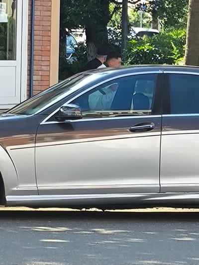 A dark-colored sedan with a light silver lower body is parked on a street. Two individuals are visible inside the car through the tinted windows. The person in the driver's seat appears to be a man with dark hair. The person in the passenger seat is partially obscured, but also appears to have dark hair. The car is parked in front of a building with a brick facade and a white door. Trees and green foliage are visible in the background, suggesting an outdoor urban or suburban setting. The street surface is paved with asphalt, and there are faint white lines visible on the road, possibly lane markers. The lighting suggests it is daytime, with sunlight casting shadows. The overall impression is a mundane scene of a car parked on a street, with people inside.