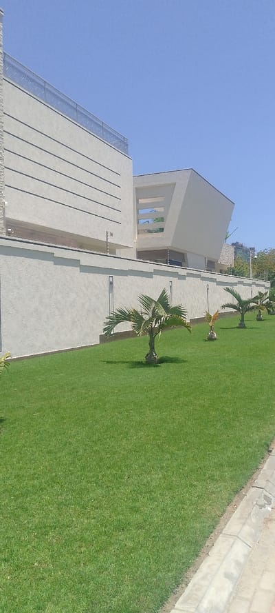 The image displays a modern residential or commercial property in Ziwa La Ng'ombe ward, Kenya, under a clear, bright blue sky. The scene is static, devoid of people or discernible activity. A large, light-colored building, featuring contemporary architectural design, occupies the background. The left section of the building has a flat facade with multiple dark horizontal lines and a metal railing along its roofline. The right section presents a more angular, geometric form with a prominent cut-out or recessed area. A textured, light-colored perimeter wall, matching the building's color, spans the midground, topped by an active electric fence with white insulators. Security cameras are mounted on the building structure above the wall. The foreground features a meticulously maintained green grass lawn. Five small, healthy palm trees with short trunks and green fronds are planted at regular intervals along the base of the wall. A grey concrete curb separates the lawn from a light-colored paved surface visible in the lower right.