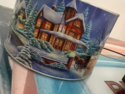 The image shows a tin decorated with a Christmas scene. The scene depicts a snow-covered house with lights on in the windows, surrounded by snow-laden evergreen trees. A horse-drawn carriage is visible in the foreground, along with a lamppost. A label on the tin partially reads "WINTER". The tin is tilted and positioned on a surface that appears to be a table or desk, with some of its reflective surface visible. The background is blurred, but shows a light-colored wall and possibly some other objects. The overall impression is of a festive, winter-themed tin, likely containing cookies or similar treats. The image does not contain any recognizable landmarks or details that would indicate its location in Balbriggan, Ireland.