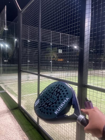 This image depicts an outdoor padel court at night, located in Privada Residencial Blue Cedar, Mexico. In the foreground, a light-skinned hand holds a dark green/teal "KUIKMA METAL" padel racket, which features a perforated face and a light purple wrist strap. The racket is positioned near a dark metal grid fence that encloses the court. Immediately beneath the fence in the foreground is a strip of artificial green turf bordering a light-colored gravel path. Beyond the fence, two padel courts with artificial green turf and white boundary lines are visible, illuminated by multiple bright floodlights. The courts are also enclosed by similar metal fencing and clear glass panels. In the background, silhouetted palm trees and faint structures are discernible against the dark night sky.