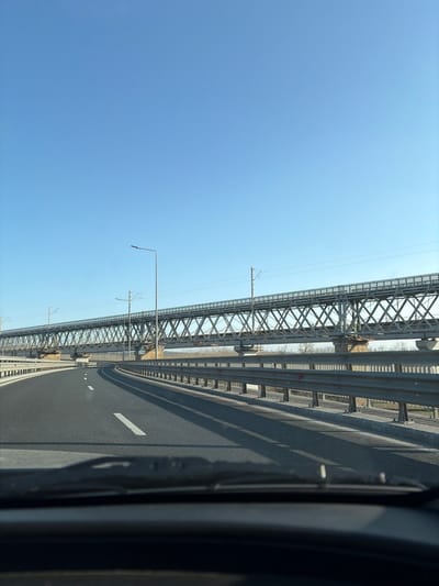 The scene, captured from a vehicle's interior, displays a multi-lane asphalt highway gently curving to the right. White dashed lane markings are present. A metal guardrail lines the right side of the highway, casting elongated shadows. A street light pole is visible on the left. In the background, the Cernavodă Bridge complex, situated near Stelnica, Romania, crosses a body of water. The complex features a prominent upper steel truss railway bridge equipped with overhead electrical lines. Below it, a secondary, lower bridge structure is supported by multiple concrete piers. Beyond the bridges, flat land with sparse vegetation is visible. The sky is clear and blue. No individuals or other active vehicles are discernable.