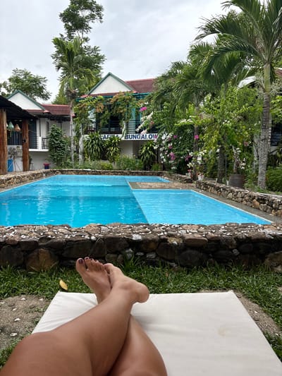 A person's legs and feet are visible in the foreground, resting on a white lounger in a tropical outdoor setting. Behind the lounger, a bright blue rectangular swimming pool with a stone border is the central focus. The pool is divided into two sections by a submerged line. Beyond the pool, a two-story building with white walls, red roofs, and balconies is visible. Lush green trees, palm trees, and flowering vines with pink and white blossoms surround the building. A sign on the building reads "CAFÉ BUNGALOW." The environment appears to be a resort or guesthouse in Bố Trạch Commune, Vietnam. The sky is overcast, suggesting a cloudy day.