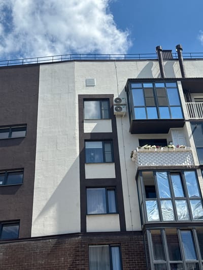 The image shows the facade of a modern multi-story building against a bright blue sky with scattered white clouds. The building's exterior features a distinct pattern of dark brown and off-white sections. Several windows of varying sizes are visible, some with dark frames and reflective glass, others with lighter frames and tinted glass.

On the right side of the building, a distinctive feature is a multi-level balcony or sunroom with large, floor-to-ceiling windows. The upper section of this feature is enclosed and appears to have blue-tinted glass panels. Below it, a smaller balcony is adorned with numerous white planters filled with flowers. A white railing is also visible on the right side of the building, extending upwards.

Two air conditioning units are mounted on the off-white section of the building, positioned side-by-side. The roofline is topped with a dark railing, and two dark, cylindrical ventilation pipes or chimneys protrude from the roof on the right side. The lower portion of the building visible in the image is constructed of dark brown brickwork.

The scene is devoid of any people or visible activity. The lighting suggests it is daytime, likely sunny, with a strong shadow cast diagonally across the off-white section of the building from an unseen structure or architectural element. The architectural style and the presence of multiple windows and balconies suggest a residential apartment building. The image is taken from a low-angle perspective, looking upwards.