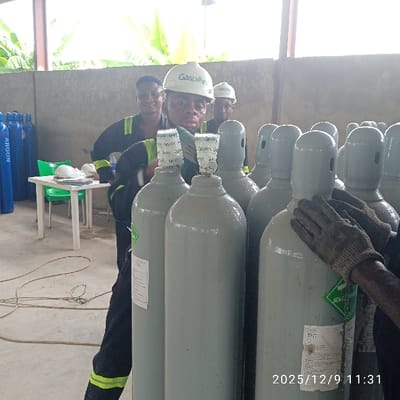 This image, taken in Pakuro, Nigeria, on December 9, 2025, at 11:31, depicts an indoor industrial setting where multiple individuals are present among numerous industrial gas cylinders.
In the foreground and mid-ground, a dense arrangement of light grey industrial gas cylinders stands upright. Many of these cylinders are fitted with caps, some secured with "GasPoint" branded tape, and several bear green diamond-shaped labels indicating "NON-FLAMMABLE GAS 2". On the far right, a pair of dark, gloved hands is seen steadying one of these grey cylinders.
Three men are visible in the mid-ground. The central figure, facing the camera with a slight smile and a visible sheen on his face, wears a dark work overall with reflective accents and a white hard hat featuring the "GasPoint" logo. Behind him to the left, another man in similar attire and a hard hat also smiles towards the camera. A third man, similarly dressed, is partially visible further back on the right.
To the far left in the background, a stack of blue gas cylinders, clearly labeled "ARGON", is visible. Adjacent to these, a small white table with two green plastic chairs is positioned. Two white hard hats, possibly identical to those worn by the men, rest on the table. Brown coiled cables or ropes lie on the concrete floor in the mid-ground.
The environment features a concrete floor and unfinished concrete block walls. Natural light floods the scene from an open side or large aperture, revealing lush green foliage, possibly banana trees, outside the structure. The overall impression is that of a gas storage or processing facility during an active work period.