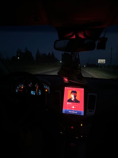 This media file captures the interior of a vehicle at night, likely in a rural setting. The dominant subject is the car's dashboard, illuminated by the glow of its screens and gauges. A large touchscreen display in the center shows what appears to be a music application with album art of a person with glasses and dark curly hair, set against an orange and red background. The track title and artist name are partially visible on the screen, but the text is too blurry to read clearly.
The dashboard features various illuminated gauges and buttons, including a speedometer and tachometer, with some warning lights showing. A hazard light symbol is visible, suggesting the car might be stopped or signaling. Outside the vehicle, the scene is dark, with hints of trees and a dimly lit road stretching into the distance. A small white sign with Arabic-like script is visible on the side of the road, an unusual detail for Belarus, where the provided location context is set. The overall mood is quiet and introspective, illuminated by the artificial light within the car, contrasting with the darkness of the night. The time of day is clearly night, and the weather appears to be calm, given the lack of visible rain or wind effects. The red glow from a light source on the left side of the frame suggests an interior car light or a light from another vehicle.