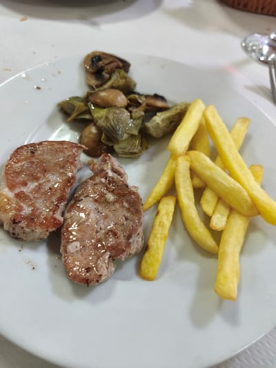 A close-up, high-angle shot captures a plate of food on a white tablecloth. The main subjects are two pieces of grilled meat, a portion of french fries, and a mix of sautéed vegetables, including artichoke hearts and button mushrooms. The meat, likely pork loin, is lightly browned with a glistening surface, seasoned with pepper. The french fries are golden and appear freshly cooked. The vegetables are tender and well-combined.
The setting appears to be an indoor dining establishment, such as a restaurant or cafeteria, in the Polígon Industrial De Picassent, Spain. The white tablecloth suggests a dining environment, and a piece of cutlery (possibly a spoon or fork) is partially visible on the right side of the plate. A blurred wicker basket-like object is faintly discernible in the upper right background. The food is presented as a complete meal, ready for consumption. No people are visible in the frame, and there is no text. The lighting is bright and even, highlighting the textures and colors of the food.