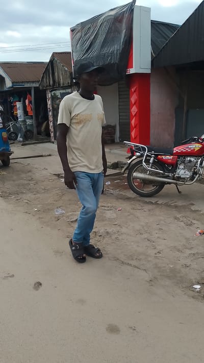 A man wearing a light-colored t-shirt, blue jeans, and black sandals walks on a sandy road in Agbarho, Nigeria. He has a dark complexion and is wearing a black baseball cap. Behind him, a red motorcycle is parked on the side of the road. To his left, a blue vehicle is partially visible. Shops with corrugated metal roofs line the street. One shop has a red facade and a black tarp covering its upper section. The ground is a mix of sand and scattered debris. The sky is overcast.