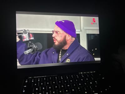 A laptop screen displays a man with a purple beanie, gold chain, and sunglasses in a recording studio. The background features the "DUHUSTLE" logo. The man is positioned behind a microphone, suggesting a podcast or interview. A Lacoste logo is visible on his jacket. Below the screen, a backlit keyboard is visible, with letters and symbols illuminated in the dark. The screen also displays a partially visible YouTube link.