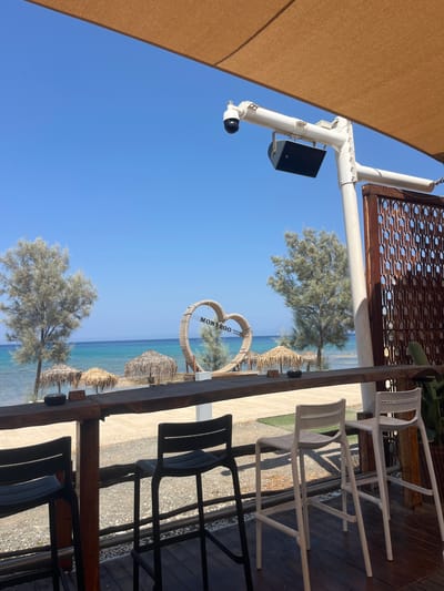 The image depicts an outdoor seating area of a establishment overlooking a beach in Protaras, Cyprus. In the foreground, a wooden bar counter is visible, with several high stools of varying colors (black and white) placed in front of it. Behind the counter, the scene opens up to a sandy beach with several thatched umbrellas and sparse trees. The turquoise sea stretches to the horizon under a clear blue sky. A decorative heart-shaped structure with the word "MONTEGO" inscribed on it stands on the beach. Overhead, a tan fabric canopy provides shade, with a security camera and a speaker mounted on a white pole structure. The overall atmosphere suggests a tranquil beachside resort or cafe.