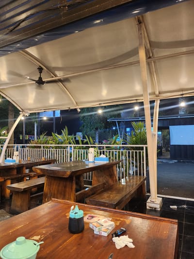 The image depicts an outdoor dining area at night, likely a restaurant or food stall, under a white tarp roof. The scene is located in Surawangi, Indonesia. In the foreground, a dark wooden table holds a light green lidded container, a small dark bottle with a turquoise cap, a pack of cigarettes, a lighter, and a crumpled white napkin. Behind this table, another similar wooden table with benches is visible. Beyond the tables, a white metal fence encloses the area, with lush green plants behind it. The background shows a dimly lit street or parking lot with trees and buildings, including a white screen on one building. Several string lights are illuminated overhead, casting a warm glow. A ceiling fan is mounted on the roof structure. The overall impression is of a casual, open-air dining establishment at night.