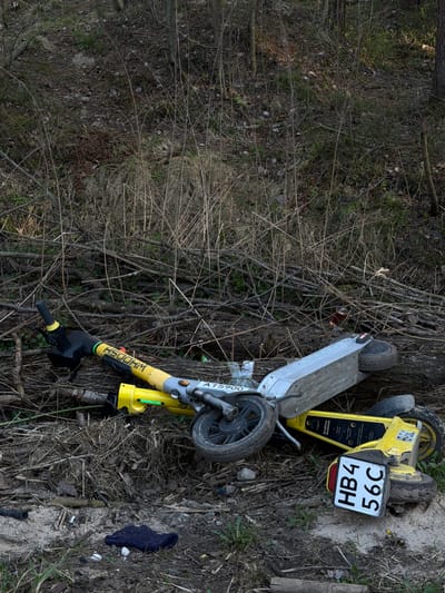 A yellow and grey electric scooter lies on its side amidst fallen branches and dirt in an outdoor setting, potentially a wooded area. The scooter has a license plate reading "HB4 56C". The brand name "WHOOSH" is visible on the scooter's handle. A dark-colored glove lies near the scooter. The location appears to be Lavriki, Russia based on the license plate and brand.