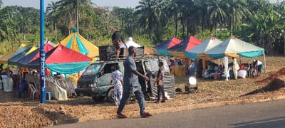 An outdoor gathering is depicted in Alayi, Nigeria, under a clear sky. Approximately six temporary canopy tents, featuring color combinations such as yellow/turquoise, red/turquoise, blue/dark gray, and white/turquoise, are erected on a dry, grassy-dirt field. Beneath these tents, numerous plastic chairs (white, blue, red) and tables covered with white cloths are arranged, accommodating roughly fifteen to twenty individuals dressed in a mix of traditional Nigerian garments and casual wear. A dark-colored panel van is parked centrally, heavily loaded with large speaker boxes on its roof rack; two individuals are on the roof manipulating this cargo. A man in a dark gray traditional Nigerian outfit and red sandals walks from left to right across the paved road in the foreground. A tall blue pole with white lettering, including "MOC," stands in the left-center near the road. A portable white satellite dish-like object is visible on a black equipment box near the tents on the right. The background features dense tropical vegetation, including palm trees and banana plants.