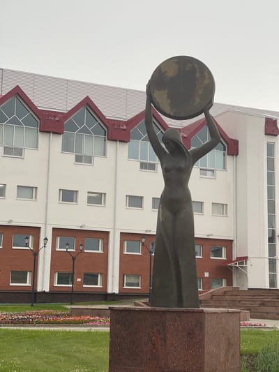 This is a vertical, eye-level, outdoor shot of a bronze statue of a woman holding a large, round disc above her head. The statue is in the foreground, to the left, and is made of dark, weathered bronze. The woman is depicted in a flowing gown, with her arms outstretched, holding the disc, which has a mottled, reflective surface.
Behind the statue is a modern building with white and red-brown brick facades. The building has many windows, some large and angular with geometric divisions, and others smaller and rectangular. The overall architectural style appears to be functional and contemporary. In front of the building and to the right of the statue, there is a neatly kept lawn with a colorful flowerbed running along its edge. Several lampposts with traditional designs are also visible in front of the brick section of the building.
The sky is overcast and a uniform light gray, suggesting a cloudy or possibly rainy day. The lighting is diffused, casting soft shadows. The scene appears to be a public space, possibly a park or plaza in front of an institutional building in Khanty-Mansiysk, Russia, given the location context. There are no people visible in the frame, and the statue's pose and the surrounding environment evoke a sense of solemnity or perhaps a tribute to an abstract concept. No text is visible in the image.