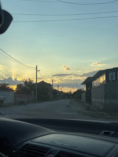 The image depicts a street scene in Belozem, Bulgaria, during what appears to be sunset or sunrise. The perspective is from inside a vehicle, looking forward. A dirt road with gravel fills the center of the frame, leading into the distance. On the right side of the road, there is a building with a dark roof and concrete walls. A sign with Cyrillic text is visible on the side of this building. The left side of the road shows houses and trees, with some appearing older and darker. Power lines crisscross the sky, silhouetted against a soft, gradient sky of pale blue and yellow hues. The sun's rays are breaking through the clouds on the left, creating a dramatic lighting effect. The foreground shows the interior of a car, including the dashboard and part of the steering wheel, indicating the image was taken from a moving vehicle or a stationary one parked on the street. The overall atmosphere is peaceful, with the natural light casting long shadows and highlighting the textures of the road and buildings.