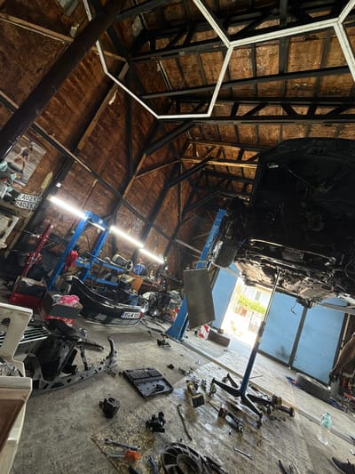 The image depicts an auto repair shop in Topolovgrad, Bulgaria. A car is suspended on a lift, with its underside exposed for inspection and repair. Various tools and car parts are scattered across the concrete floor, suggesting ongoing work. The workshop has a rustic, wooden-paneled ceiling and is illuminated by fluorescent lights. In the background, a large blue garage door is open, revealing a sunlit exterior. A person's arm is visible on the right side of the frame, indicating their presence in the workspace. The overall scene conveys a busy and functional environment for automotive maintenance.
