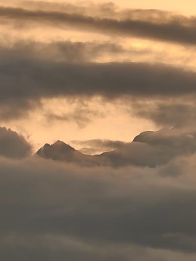 The image depicts a dramatic cloudscape with mountain peaks piercing through the fog. The sky is a blend of golden hues and dark, ominous clouds, suggesting either sunrise or sunset. The mountain peaks are silhouetted against the brighter sky, creating a stark contrast with the surrounding mist. The overall impression is one of a remote and rugged landscape, possibly an alpine region. The image does not show any elements that would indicate its location in Aleo, India.