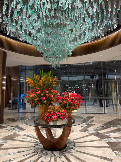 The image shows the interior of the NUSTAR Resort and Casino in Cebu City, Philippines. The scene is dominated by a large, elaborate chandelier made of numerous translucent, pale green, leaf-shaped glass pieces hanging from the ceiling. Below the chandelier, a glass-topped circular table with a wooden base is set on a patterned marble floor. On the table are two large floral arrangements. The arrangement in the foreground is composed of bright red anthurium flowers and smaller yellow and orange blooms, in a woven basket. To the left and slightly behind it, a second, larger arrangement features vibrant orange and yellow heliconia flowers mixed with red anthuriums and other greenery, also in a woven basket.
In the background, a glass facade reveals the interior of what appears to be a casino, with reflections of lights and possibly signage reading "CASINO". Several people are visible through the glass, some walking and others standing. The floor is a complex mosaic of marble tiles in shades of beige, brown, and grey, with a radiating sunburst pattern. The overall impression is one of a luxurious and modern lobby.