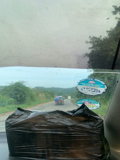 An exterior, eye-level, medium shot shows a view from inside a vehicle looking forward through the windshield. The windshield is dirty and scratched, with a black plastic bag obscuring the lower part of the view. Outside, a road curves through a lush, green landscape with trees and hills under a cloudy sky. A few vehicles are visible on the road, including one with people in the back. Two stickers are visible on the windshield: one at the top reads "NATIONAL UNION OF ROAD TRANSPORT WORKERS 2505 VALID FOR YEAR", and a second one below it has similar text and a logo. The location is Assob Hausa, Nigeria.