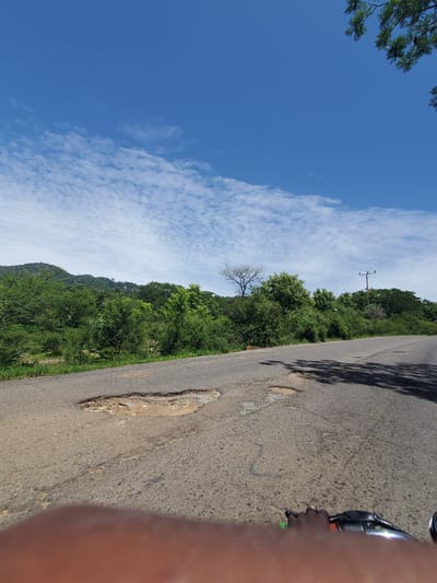 The image depicts a road with several potholes, suggesting poor road conditions. The road curves to the right, leading into a landscape with lush green trees and hills in the background under a bright blue sky with scattered white clouds. A telephone pole with wires is visible on the right side of the image. In the foreground, a person's hand and part of a motorcycle handlebar are visible, indicating the photo was taken from a moving vehicle. The scene is likely a rural or semi-rural area, possibly on the outskirts of Gwolong, Nigeria, given the presence of natural landscape and the general appearance of the road and infrastructure. There are no other individuals or significant interactions visible in the frame.