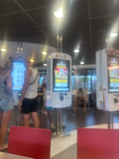The image depicts a restaurant interior, likely a fast-food establishment, with multiple self-ordering kiosks visible. The scene is vertically oriented. In the foreground, a person is standing in front of a kiosk, their arm extended as if interacting with the screen. Another person is partially visible in the background, also near a kiosk. The kiosks feature illuminated screens displaying what appears to be a menu with images of food items. The setting includes tables and chairs, with red-colored surfaces prominent on the right side of the frame, possibly tabletops or menu boards. The lighting suggests an indoor environment, possibly during daylight hours given the brightness. There are no discernible weather cues. The overall atmosphere seems casual, typical of a quick-service restaurant. No specific text is clearly legible on the kiosks or in the environment. The location is identified as Narbonne, France.
