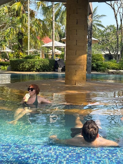 A woman in a green swimsuit and sunglasses is drinking from a cup in a pool, with a man partially submerged in the same pool. The pool is bordered by blue tiles on one side and a textured stone structure on the other. Lush green trees and vegetation surround the pool, and the sky is visible in the background. This location appears to be in Khok Kloi, Thailand.