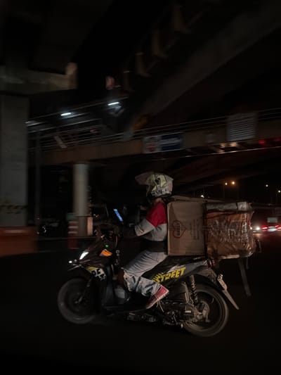 A delivery driver on a motorcycle is parked on a Pasay City, Philippines street. He is wearing a helmet, a red and grey sweatshirt, and denim jeans with red shoes. He is sitting on his motorcycle, which has a delivery box strapped to the back. The box is labeled "Geely". The motorcycle is parked next to a building with a metal staircase. The environment is dark, illuminated by street lights and the motorcycle's headlight.