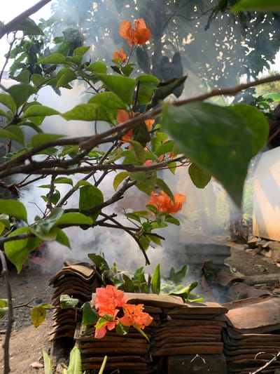 The image shows a close-up view of orange bougainvillea flowers and green leaves in the foreground, with smoke rising in the background. The smoke appears to be coming from a pile of terracotta roof tiles stacked on the ground. The scene is outdoors, with trees and foliage visible behind the smoke. The lighting suggests it is daytime, possibly morning or late afternoon. The overall impression is one of a rustic, possibly rural, setting in Karangkemiri, Indonesia, where traditional practices like controlled burning or cooking might be taking place, evidenced by the smoke and the stacked roof tiles. There are no people visible in the frame.