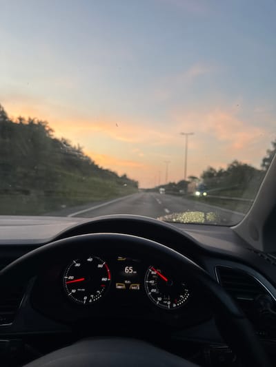 The image depicts the interior of a vehicle, looking forward through the windshield at a highway during sunset. The sky exhibits a gradient of blue to pale orange, with hints of pink clouds. Trees line the left side of the road, which recedes into the distance. Streetlights are visible on the right side of the highway.
In the foreground, the steering wheel and dashboard are prominent. The speedometer indicates a speed of 65 mph. A digital display shows "16.0k" and "149917" and "9457.8". The vehicle is traveling on a paved road, likely a motorway, given the presence of multiple lanes and streetlights.
While the specific location within the Borough of Swale, United Kingdom, cannot be definitively determined from the visual information alone, the presence of a motorway, road infrastructure typical of the UK, and the overall landscape suggest a possible setting within that region. There are no people visible in the image, nor any specific landmarks that would pinpoint the exact location. The scene captures a moment of travel during twilight hours.