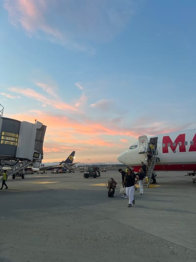 The image depicts an airport scene at sunset in Ferno, Italy, likely at Milan Malpensa Airport. In the foreground, passengers are disembarking from a white airplane with large red lettering that appears to spell "MIA". Some passengers are descending a set of stairs while others are walking on the tarmac. To the left, a jet bridge is connected to another airplane, a Ryanair aircraft, with the number "609" visible. Several service vehicles, including baggage carts and a small utility vehicle, are present on the tarmac. The sky is filled with soft pink and orange hues from the setting sun, contrasting with the clear blue sky. The overall atmosphere is one of travel and departure or arrival during twilight.
