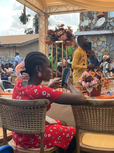 An outdoor social gathering is depicted in Kumasi, Ghana, under a light-colored, draped canopy. In the immediate foreground, a woman with braided hair, adorned with a pink scrunchie and red floral earrings, is seated in a wicker-backed chair. She wears a red short-sleeved dress with a white and blue floral pattern. She holds a black smartphone horizontally with both hands, viewing its screen.
Adjacent to her, a table is set with an orange tablecloth, featuring a floral centerpiece of burgundy, orange, and cream-colored artificial flowers on a golden stand. A clear wine glass and a plastic bottle of Voltic water are visible on the table.
In the mid-ground, slightly out of focus, a bearded man stands, facing right, wearing a distinctive yellow long-sleeved tunic with black patterns. He holds a dark object, possibly a phone, in his left hand. Other guests are seated and standing throughout the background, largely out of focus.
The background features residential architecture: a building with a textured stone facade and three discolored satellite dishes mounted on its exterior, and the roof of another building with light-colored corrugated sheeting. The sky is partly cloudy.