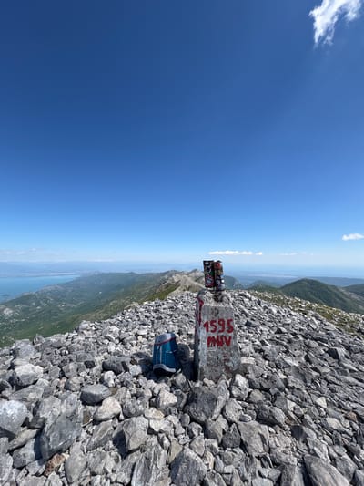 The image shows a mountain peak in Mali Mikulići, Montenegro. A concrete pillar, painted with "1595 MNV" in red, stands amongst a rocky terrain. The pillar also has several stickers attached to its top. A blue backpack rests against the pillar. In the background, the sky is clear blue, with a distant view of mountains and a body of water.