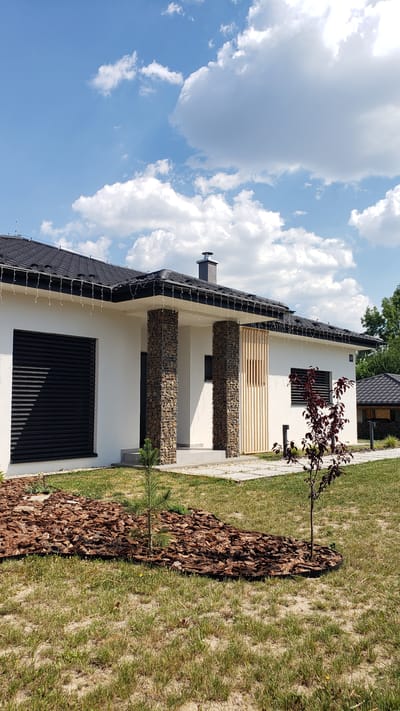 The image captures a modern house with a unique architectural design, featuring white walls, a dark-tiled roof, and prominent gabion pillars filled with stones. These pillars support a covered patio area, which is further accented by vertical wooden slats. The house has dark-colored window shutters and what appears to be outdoor string lights running along the eaves.
In front of the house, a lawn area is partially covered with mulch, forming a decorative border. Two young trees are planted in this mulched area. The larger tree, with dark red leaves, stands closer to the house, while a small pine sapling is visible near the bottom of the frame. The setting is a residential area, likely a backyard, given the presence of the house and manicured landscaping.
The sky is bright blue with fluffy white clouds, suggesting a clear and sunny day. The time of day appears to be daytime, likely late morning or early afternoon, given the lighting and shadows. The image is oriented vertically, with the house and lawn filling the majority of the frame. There is no visible text in the image. The overall impression is one of a well-maintained, contemporary home in a suburban or semi-rural setting.