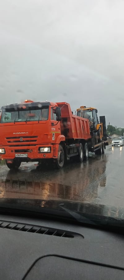 An orange KAMAZ dump truck is transporting a yellow backhoe loader on a flatbed trailer. The truck has its hazard lights on, and the backhoe is partially covered by the truck's dump body. The scene is on a wet road, indicated by reflections on the asphalt, suggesting it is raining or has recently rained. The sky is overcast and gray, implying a cloudy or stormy day. In the background, other vehicles, including a white car, are visible on the road, along with some trees and buildings suggesting an urban or semi-urban environment. The image appears to be taken from inside another vehicle, looking out through the windshield, which has some streaks or water droplets.

Visible text includes "KAMAZ" on the front of the truck and a license plate that reads "K 960 HH 716". There is also some Cyrillic text on the side of the truck and on the backhoe, but it is not clearly legible. The overall mood is functional, with the focus on the transportation of construction equipment on a challenging weather day. The location context provided is Kabanikha, Russia.