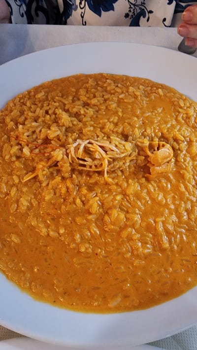 The image shows a close-up of a plate of seafood risotto, likely at a restaurant in Latina, Italy. The risotto is a vibrant orange color and appears to be creamy, with individual grains of rice visible. In the center of the dish, there are pieces of seafood, including what looks like a small crab leg and some shredded white meat. A single red thread, possibly saffron, is visible on top of the seafood. The plate is white and rests on a white tablecloth. In the blurred background, a person's hand with painted nails is visible, suggesting someone is about to eat the dish or has just finished. The context of Latina, Italy, implies the dish is likely prepared with local ingredients and follows regional culinary traditions.