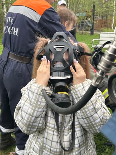 In Grebnevo, Russia, a person wearing a plaid jacket is shown putting on a black respirator mask with a black hose and yellow filter. The individual's hands are visible on the mask, adorned with light-colored floral nail art. To their left, a person wearing a dark blue uniform with orange and silver accents stands close. The uniform has white text visible on the left side. In the background, there is green grass, a tree, and a piece of silver equipment.
