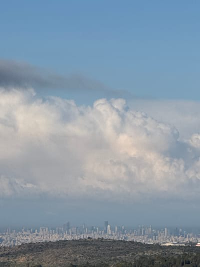 An elevated long-distance view from the city of Rosh Haayin, Israel, captures a panoramic landscape. The upper third of the image features a bright, clear blue sky. Below this, a dense layer of white to light grey cumulus clouds extends horizontally across the middle of the frame, with some darker grey cloud formations visible on the upper left. Directly beneath the clouds, a hazy urban skyline stretches across the horizon, dominated by numerous high-rise buildings and skyscrapers, indicative of a large metropolitan area in the distance. The lowest portion of the image shows dark, undulating hilly terrain covered in dense, low-lying vegetation, likely trees or shrubs. No people or specific actions are discernible. The scene is static, depicting a wide vista under a partially cloudy sky.