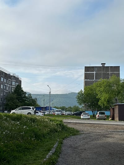 A street scene in Vilyuchinsk, Russia, shows a gravel road in the foreground curving to the left. To the left of the road is a patch of grass with dandelion seed heads and green foliage. Further left, a large, multi-story apartment building with a concrete facade and visible balconies or window frames dominates the scene. A white minivan and other cars are parked along the side of the road in front of the building. In the middle ground, a taller, square concrete building with a dark rooftop structure stands prominently. Trees with green foliage are situated in front of this building. Further back, a blue structure is visible. In the background, a hazy, green-hilled landscape under a cloudy sky forms the backdrop. Several cars are parked in a lot between the buildings and the distant hills. A street light is visible near the center of the image. The sky is mostly overcast with patches of blue.